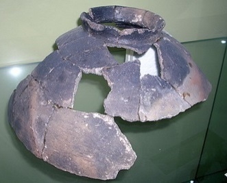 Na slici se vidi ostatak glinene posude, koju čine mnogo zasebnih dijelova koji su zalijepljeni međusobno. Posuda ima oblik polovice okrugle vaze sa užim otvorom na vrhu. Predmet stoji na staklenoj podlozi.